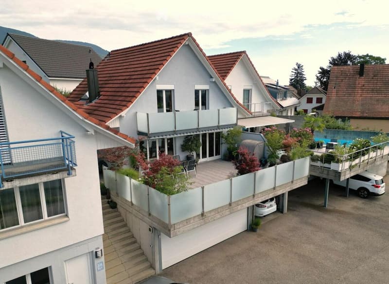 A modern two-story house with red-tiled roofs, large windows, and multiple balconies. The house features a raised ground floor with a covered parking area and a small garden area with potted plants and outdoor furniture.