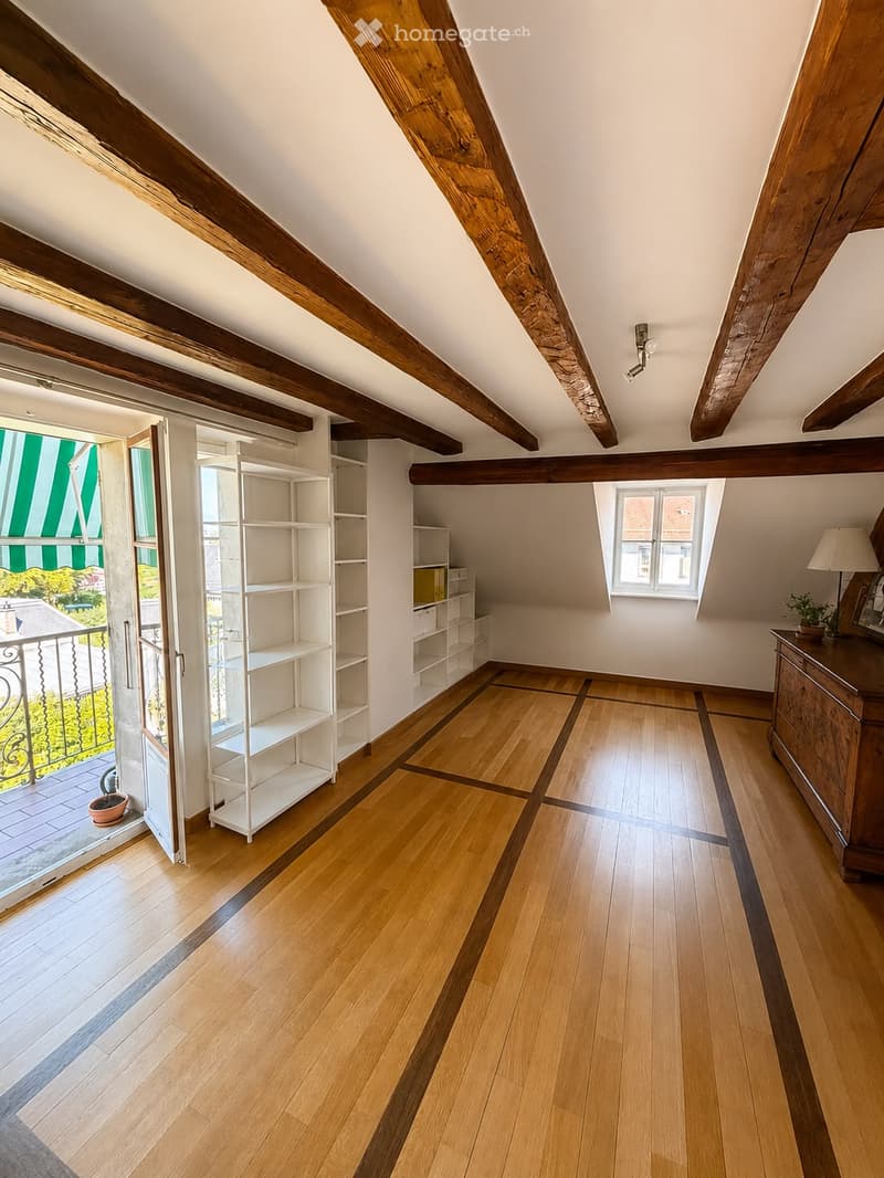Bright attic room with wooden beams, wooden flooring, white shelving unit, small table with lamp and picture frame, and a view to a balcony.