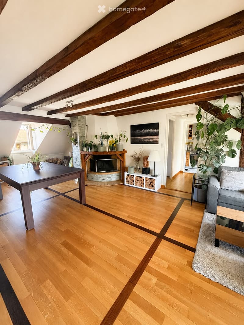 Bright living room with wooden floors, exposed wooden beams, a fireplace, a sofa, a dining table, and several plants.