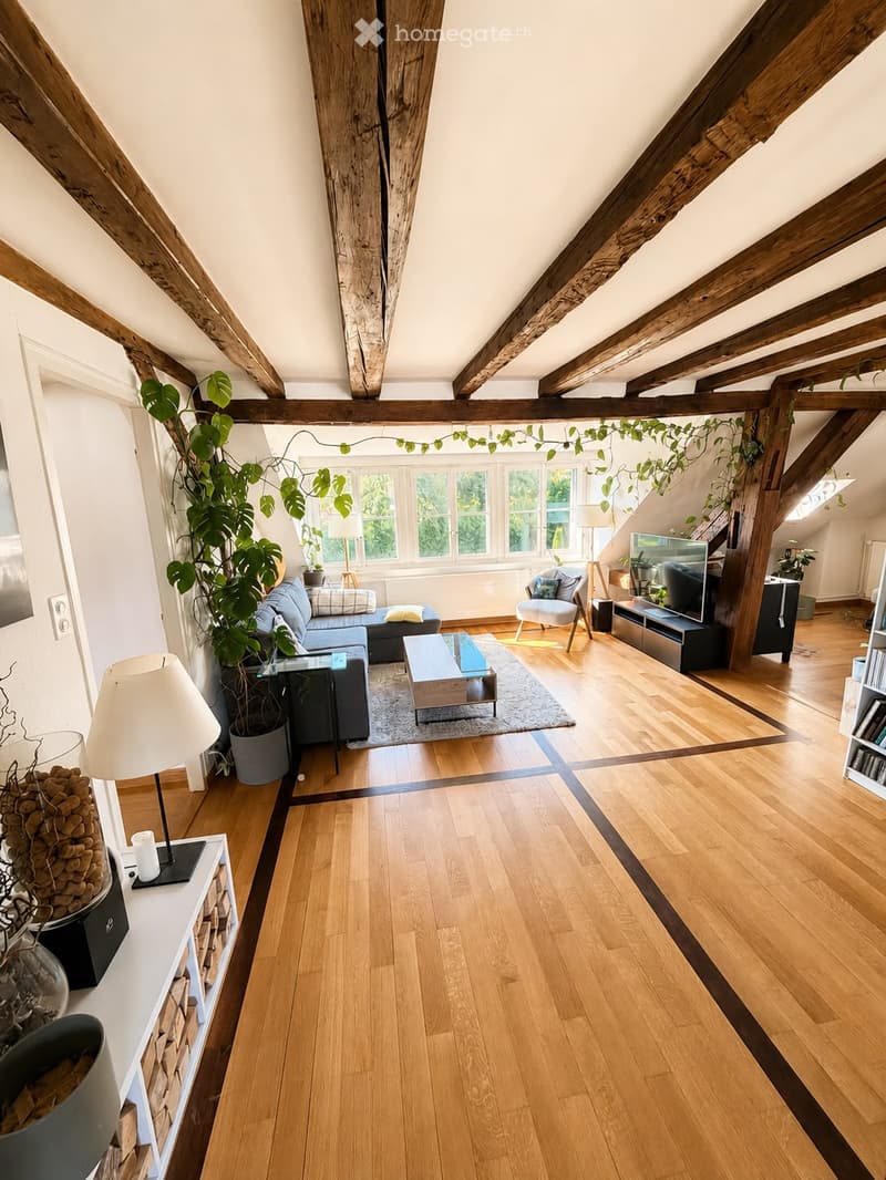 Bright living room with wooden floors, exposed wooden beams, large windows, sofa, coffee table, armchair, table lamp, television, bookshelf, and potted plants.