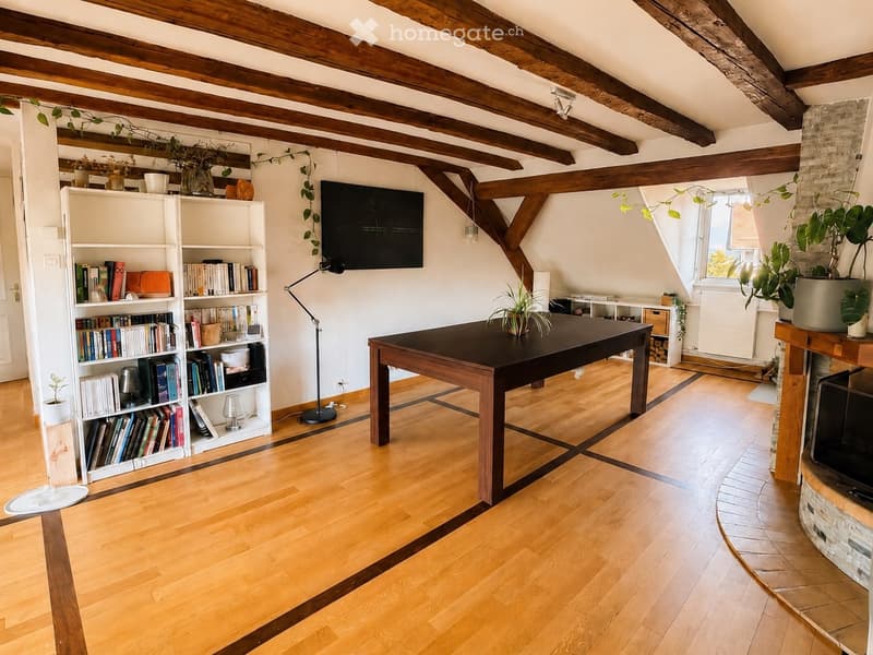 Bright living room with wooden floors, exposed wooden beams, a large table, bookshelf, TV, floor lamp, and fireplace.