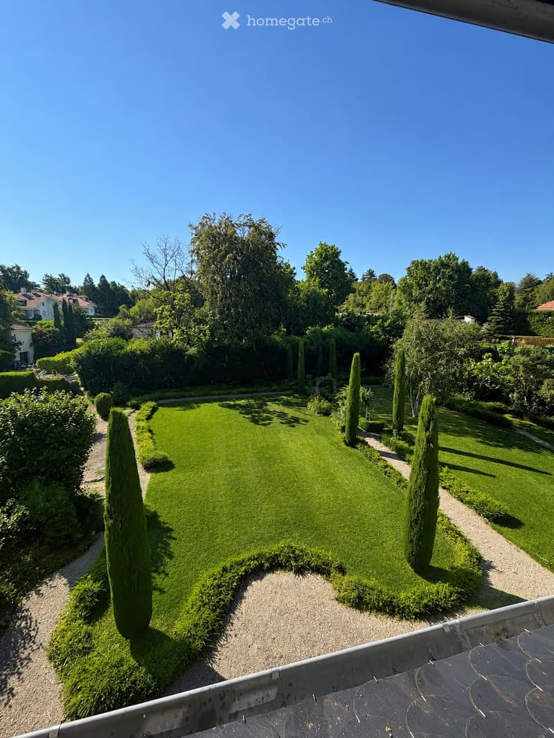 Well-maintained garden with a central lawn, neatly trimmed hedges, and tall coniferous trees lining the pathways.