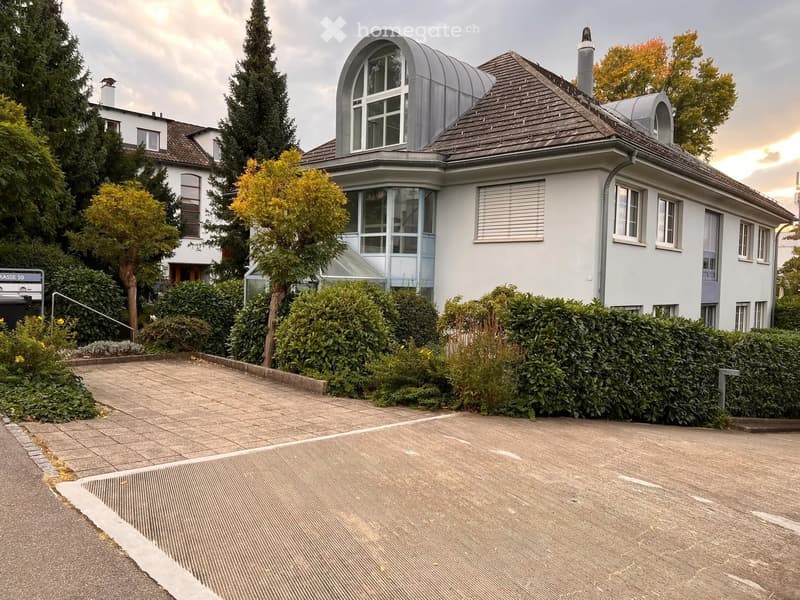 Modern two-story house with a grey roof, large windows, and a well-maintained garden with hedges and trees.