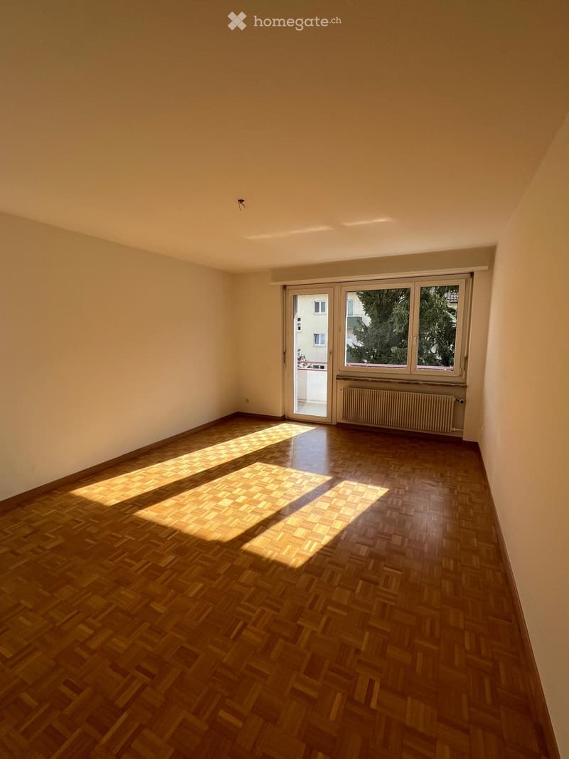 Empty room with wooden parquet flooring, white walls, and large windows with blinds. Natural light streams in, casting shadows on the floor.