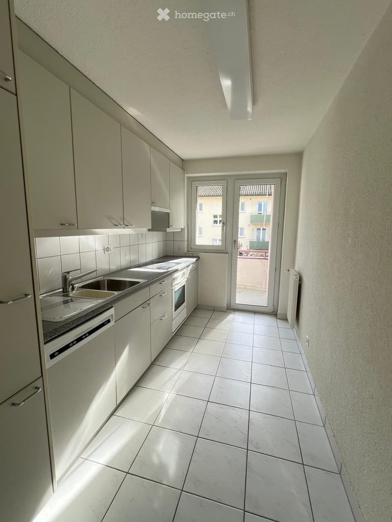 Modern kitchen with white tiled floor, white cabinets, stainless steel sink, oven, and dishwasher. Features a sliding glass door leading to a balcony.