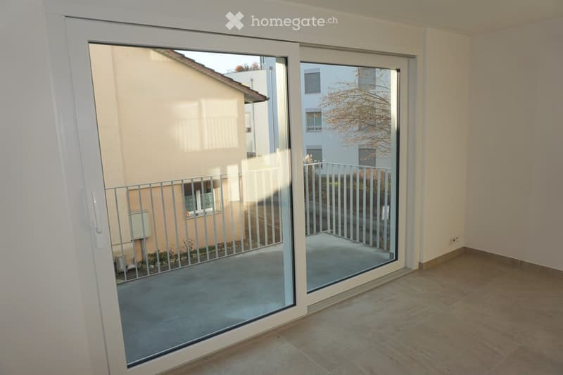 View of a modern balcony area with large sliding glass doors, tiled floor, and metal railing, overlooking neighboring buildings and a tree.