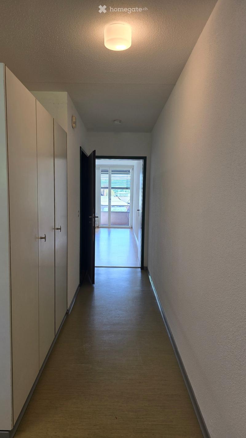 A narrow hallway with wooden flooring, white walls, and a single ceiling light. An open door at the end leads to a balcony or outdoor area.