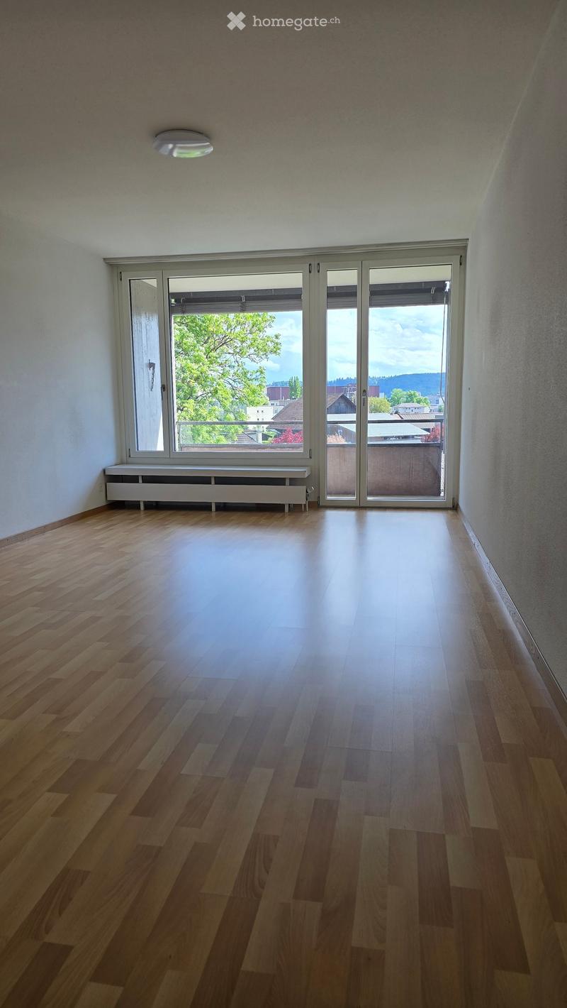 Empty room with wooden flooring, white walls, large sliding glass doors leading to a balcony, and a small white bench against the wall.