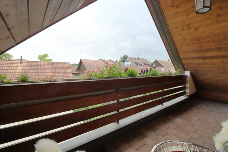 Wooden balcony with a wooden railing, tiled floor, and potted plants on the railing. The background shows a residential area with houses and greenery.
