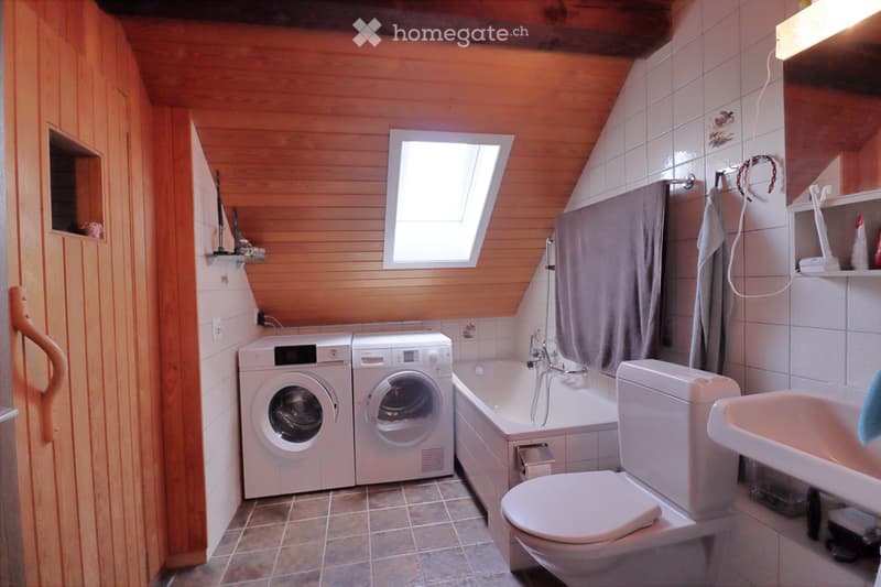 Bathroom with wooden walls, tiled floor, skylight window, washing machine, dryer, bathtub, toilet, sink, and shelves with towels and toiletries.