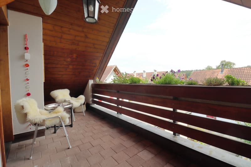 Balcony with wooden railing, brick floor, two fur-covered chairs, small table, and decorative wind chime.