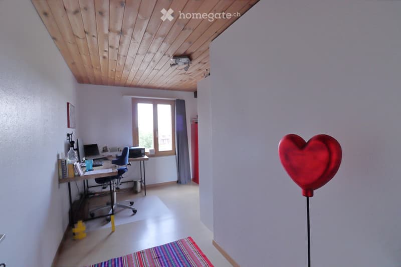 Bright room with wooden slanted ceiling, white walls, large windows, desk with laptop, chair, printer, books, and a red heart-shaped balloon on a stand.