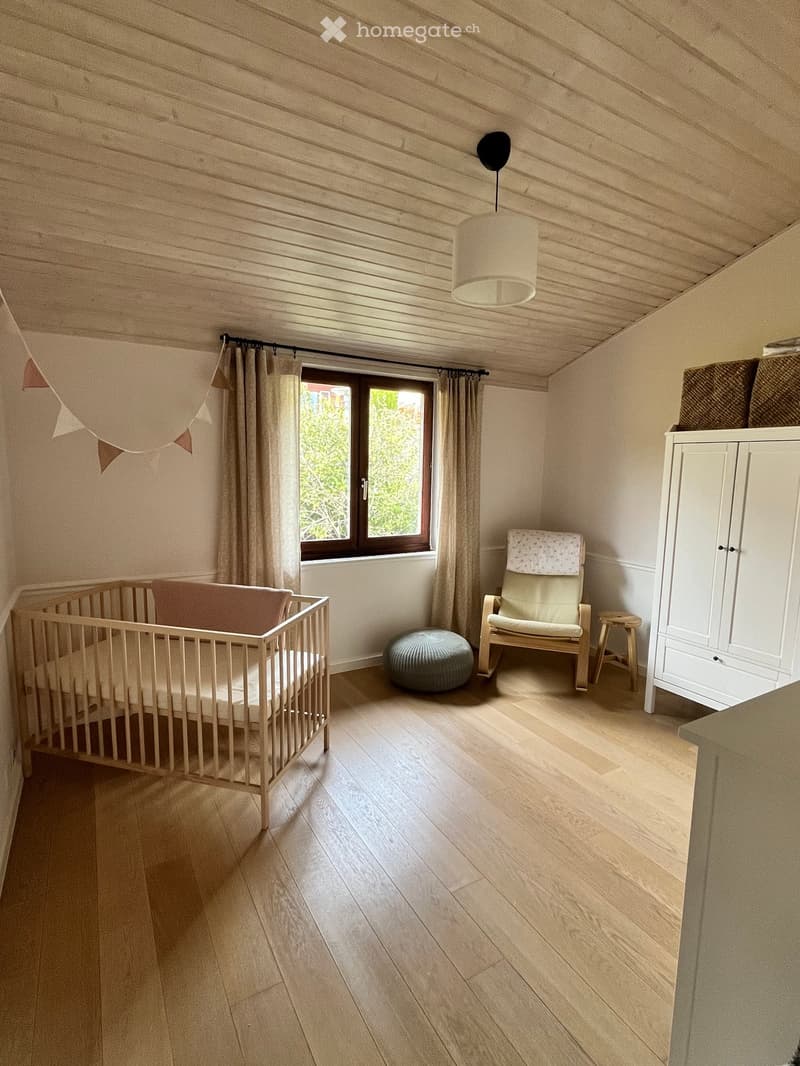 Bright, cozy bedroom with wooden flooring, a crib, a white cabinet, a hanging pendant light, and a window with curtains.