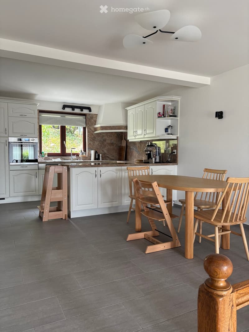 Modern kitchen with white cabinets, grey tiled floor, wooden dining table with chairs, and built-in appliances including an oven and fridge.