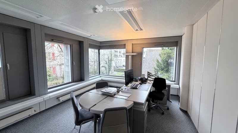 Modern office space with large windows, a round table with chairs, a computer workstation, and grey cabinetry.