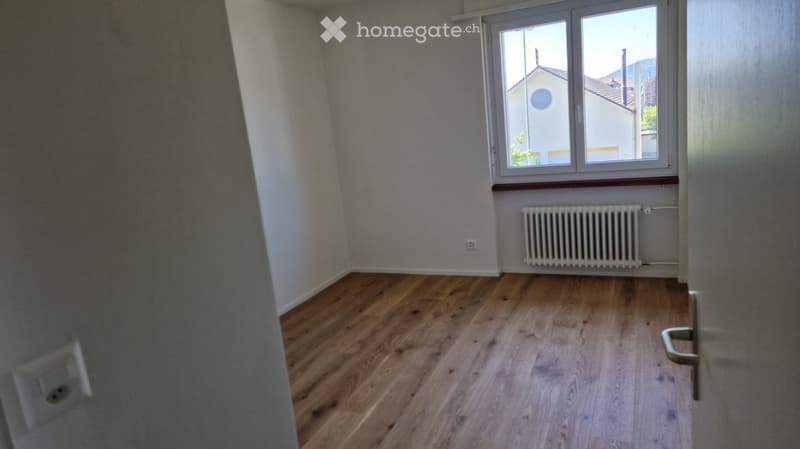 Empty room with wooden flooring, white walls, a window with a view of a building, and a radiator below the window.