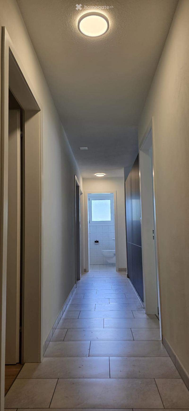 A clean, well-lit hallway with beige tiled flooring, white walls, and recessed lighting. Doors lead to various rooms, including a visible bathroom at the end.