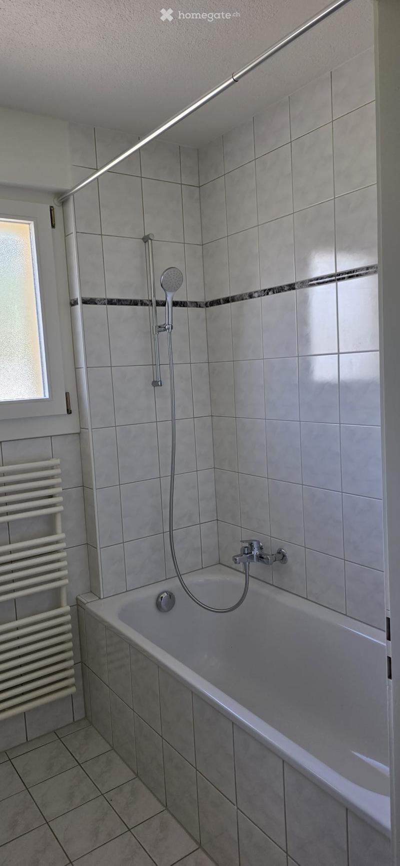 Modern bathroom with a bathtub, showerhead, faucet, and radiator. The walls are tiled in light grey with a decorative border, and the floor is tiled in a matching pattern.