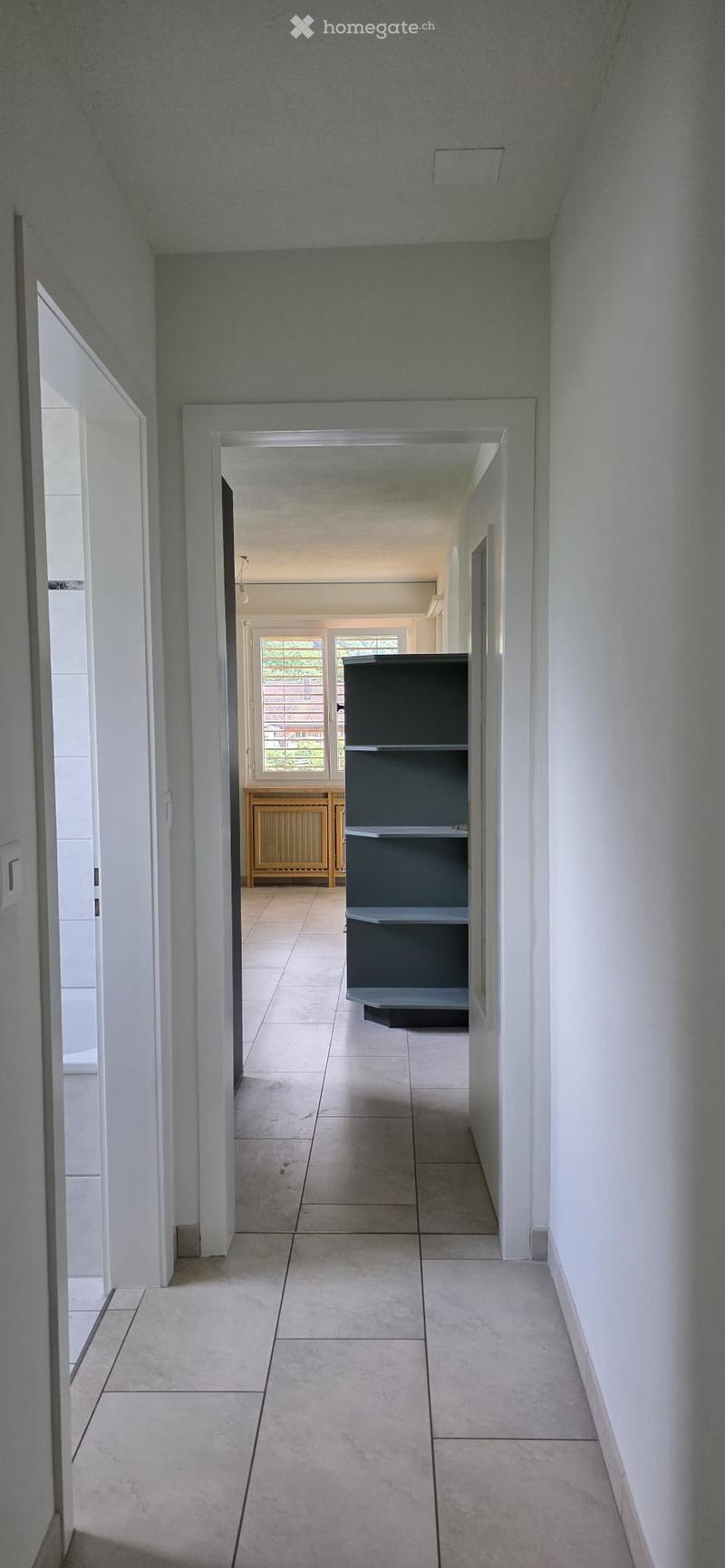 A clean, modern hallway with tiled flooring and white walls, featuring a grey shelving unit and a glimpse of a room with a radiator and window in the background.