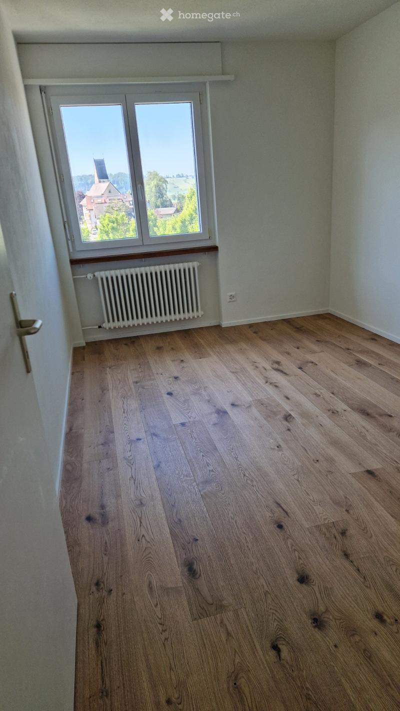 Empty room with wooden flooring, white walls, a radiator, and a large window offering a view of the countryside.