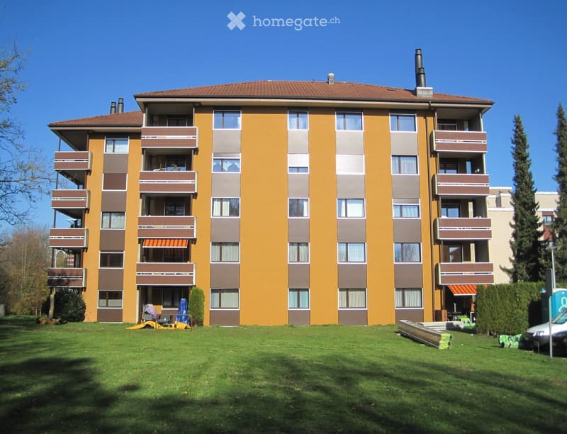 Four-story residential building with yellow facade, brown balconies, and multiple windows. The building has a well-maintained lawn in front with some outdoor furniture and construction materials visible.