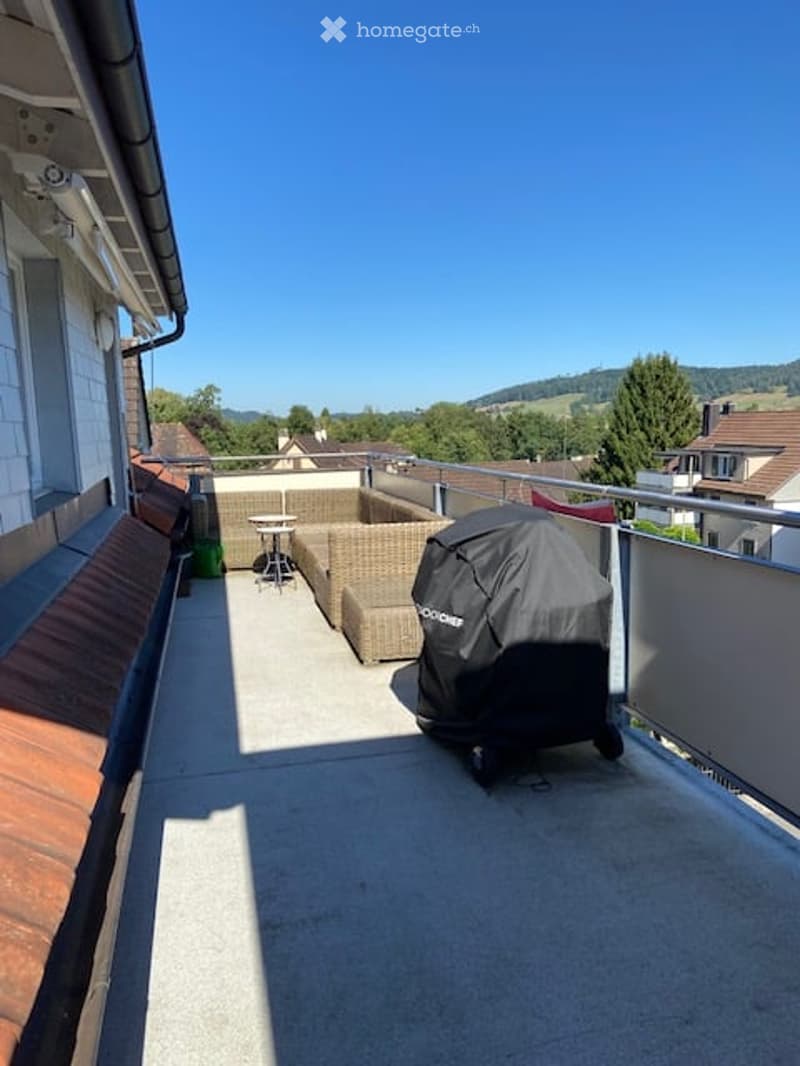 Balcony with a covered grill, outdoor seating area including a sofa and a small table, overlooking a green landscape.