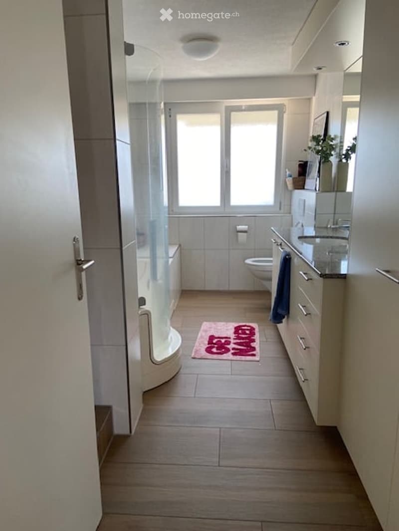 Modern bathroom with a bathtub, toilet, sink, and mirror. The space features light-colored tiles, a wooden floor, and a small pink rug with text on it.