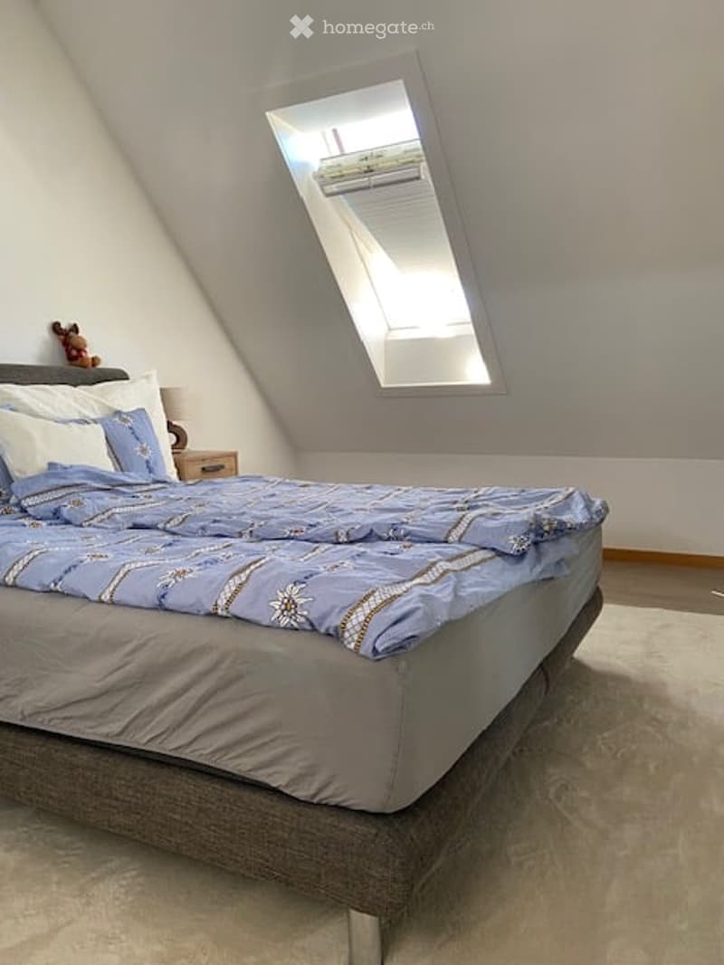 Cozy attic bedroom with a large window, a neatly made bed with blue and white bedding, a nightstand with a lamp, and a small stuffed toy on the nightstand.