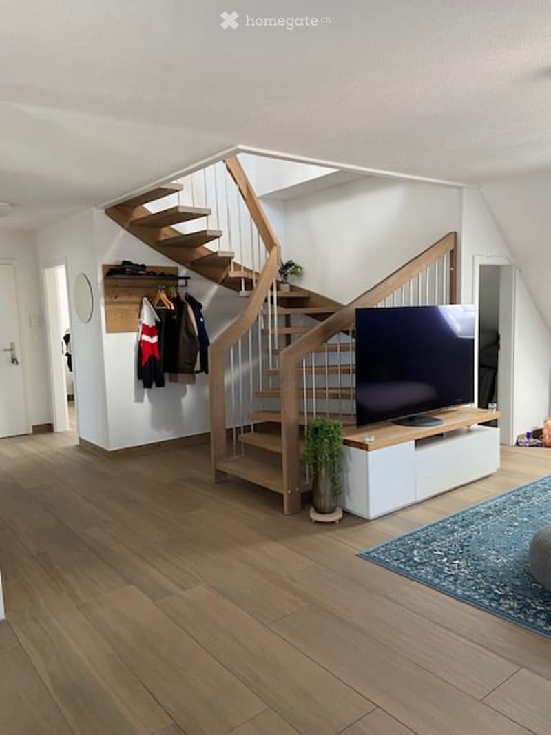 Modern living area with wooden flooring, a staircase leading upstairs, a flat-screen TV on a white stand, a coat rack with jackets, and a small potted plant near the stairs.