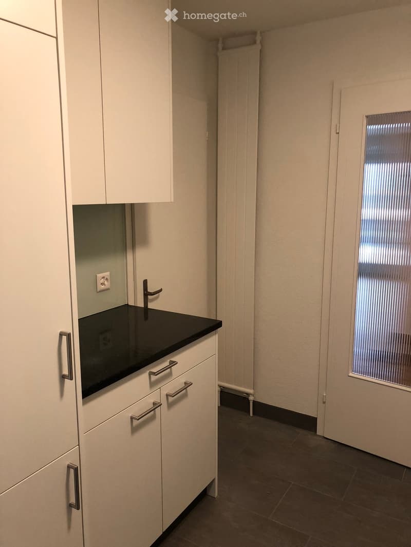 Empty kitchen area with white cabinets, black countertop, and a window with blinds. No appliances or furniture are present.