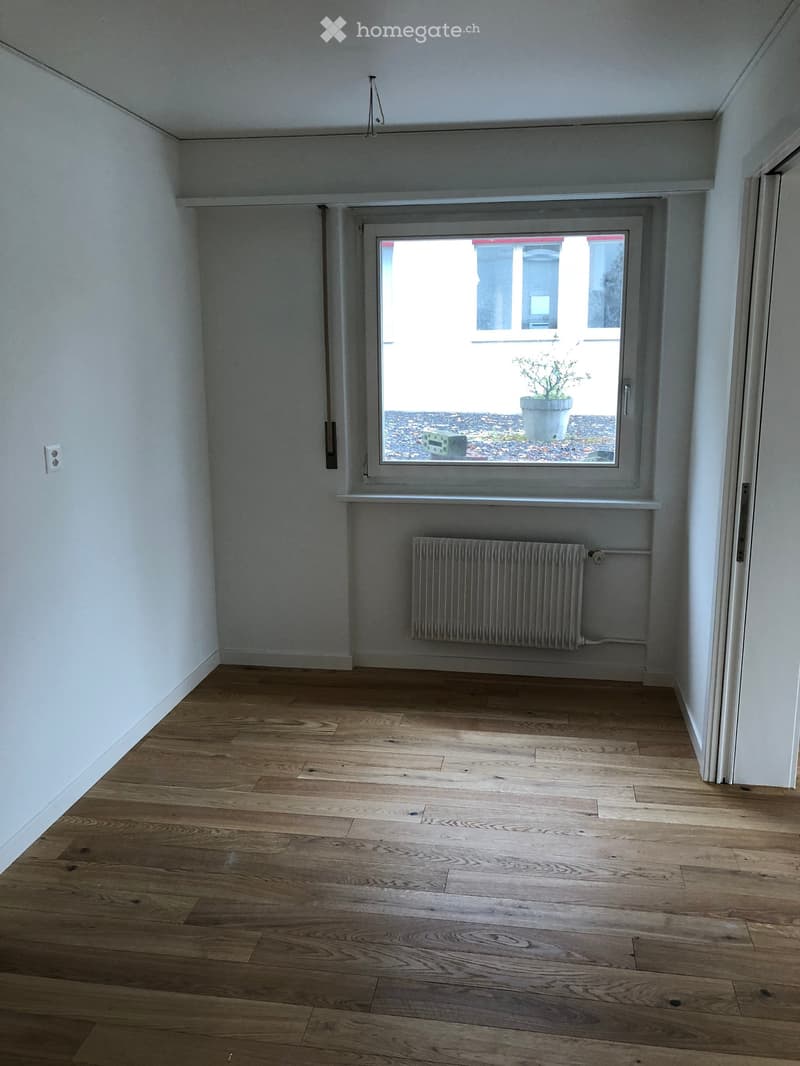 Empty room with wooden flooring, white walls, a window with a radiator below, and a visible electrical outlet on the wall.