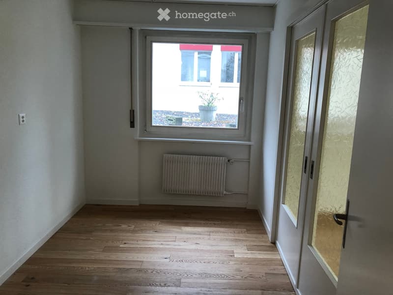 Empty hallway with wooden flooring, white walls, a radiator, and a frosted glass door leading to an outdoor area.