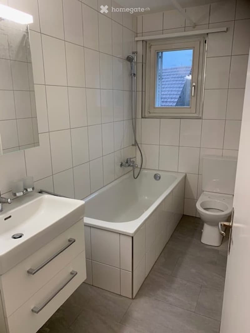 Modern bathroom with white tiles, a bathtub, toilet, sink, and a window with a view of a roof.