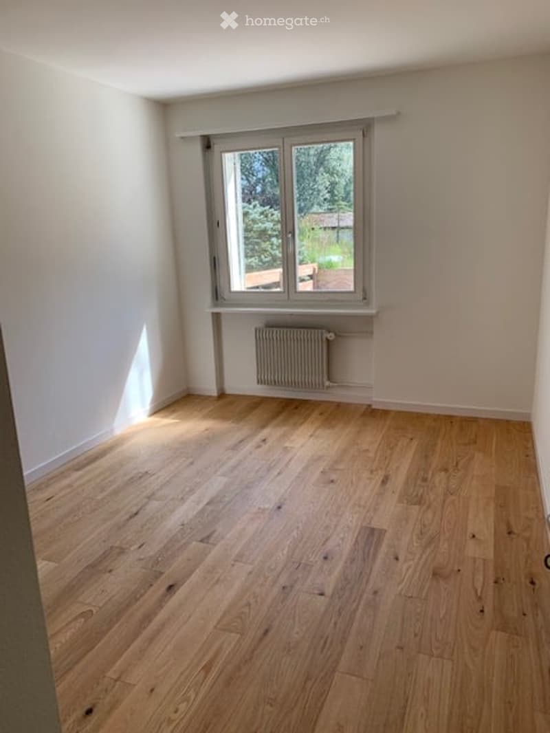 Empty room with light wooden flooring, white walls, and a single window with a radiator below it.