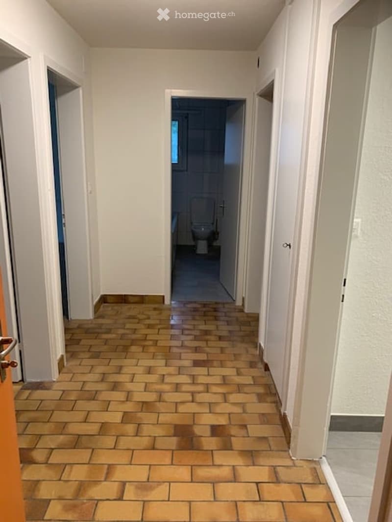 Empty hallway with tiled floor and white walls, leading to a bathroom at the end.