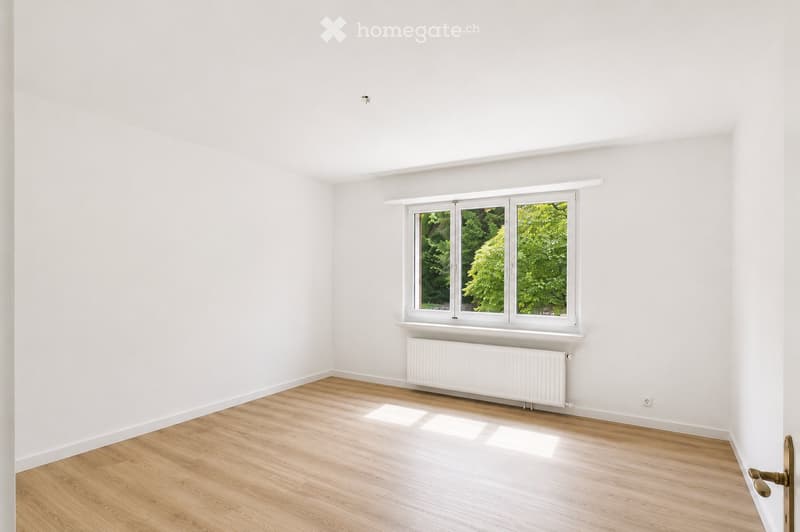 Empty room with white walls, wooden flooring, and a large window with white frame showing greenery outside. A white radiator is placed below the window.