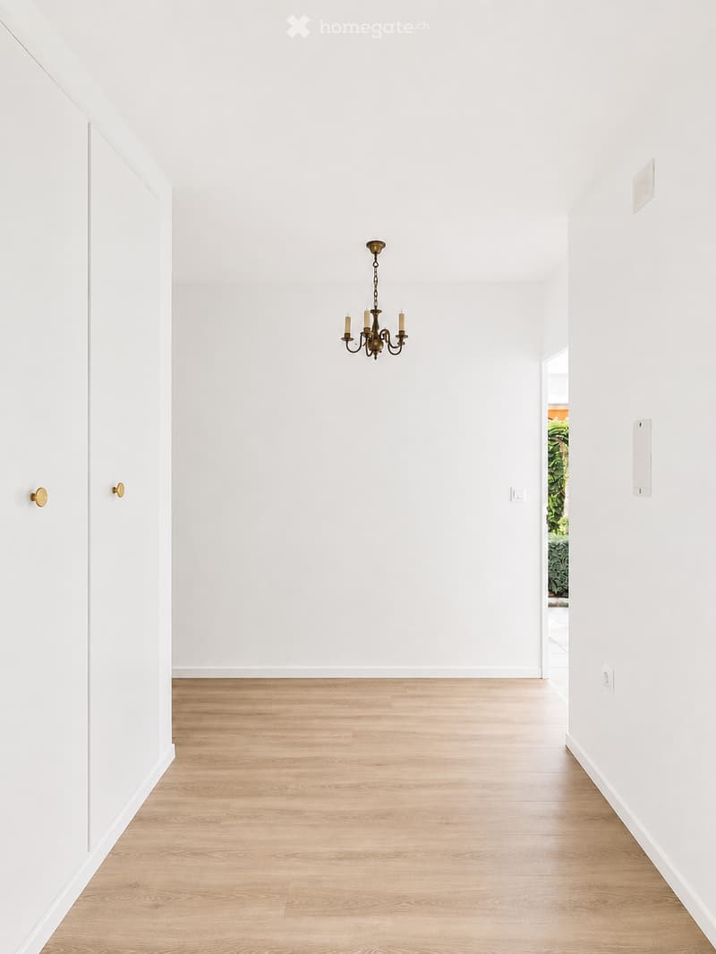 Empty hallway with white walls, light wooden flooring, and a simple chandelier with four candles.