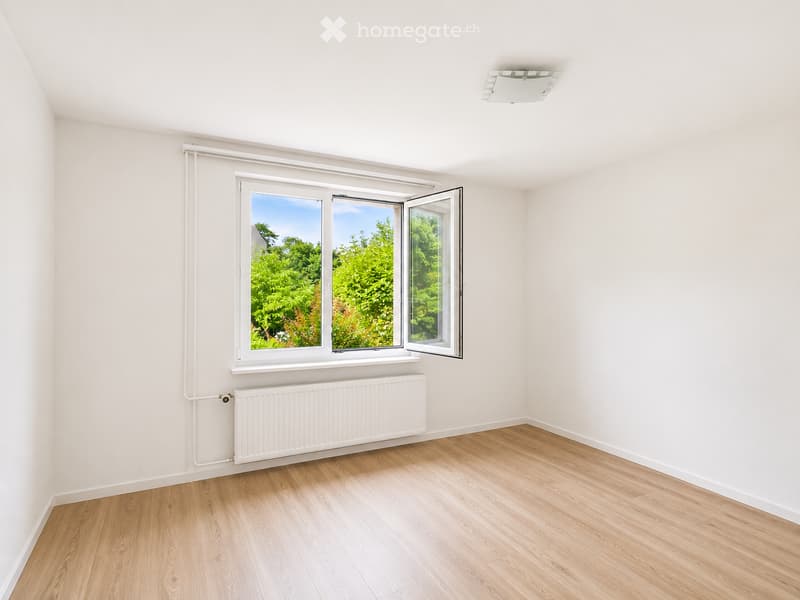 Empty room with light wooden flooring, white walls, a large window with white frame, and a white radiator below the window.