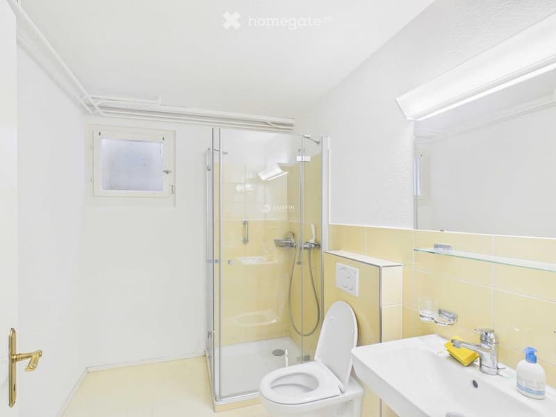 Modern bathroom with a glass-enclosed shower, toilet, sink, and mirror. Features include a window, tiled walls, and a glass shelf above the sink.