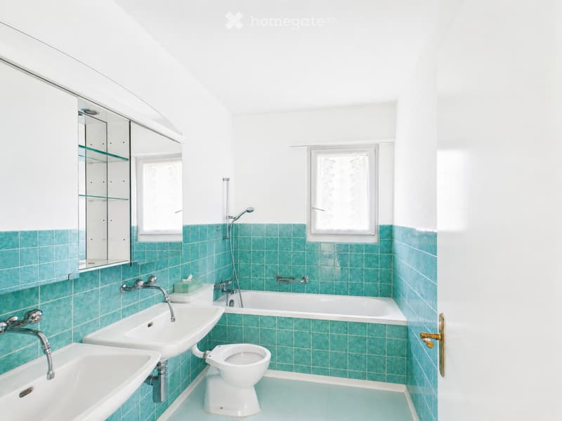 Bright bathroom with teal tiles, white fixtures, and two windows. Includes a sink, bathtub, toilet, and a mirrored cabinet.