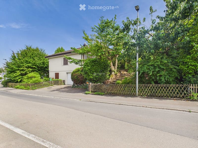 A two-story house with a beige exterior, surrounded by greenery and a wooden fence. The house has a driveway and is situated on a quiet street.