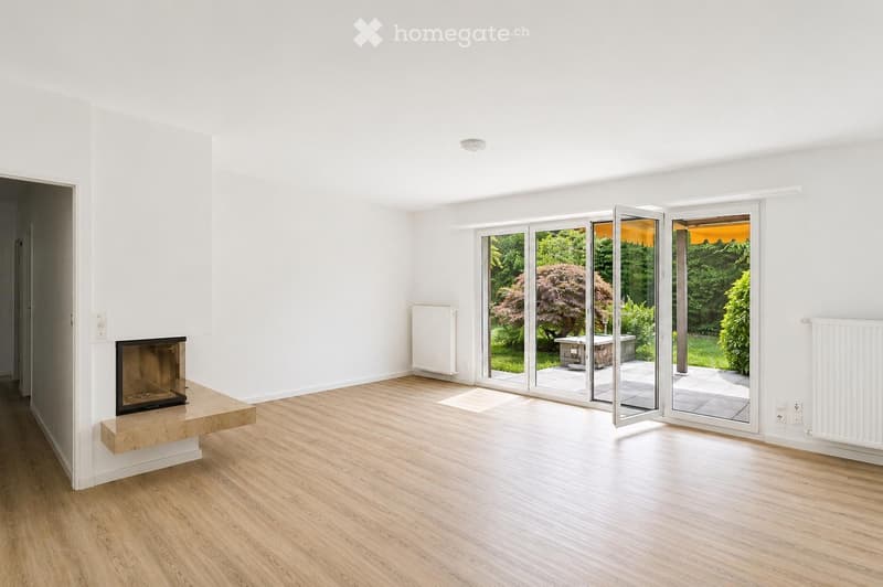 Bright, unfurnished living room with white walls, wooden flooring, fireplace, large sliding glass doors opening to a garden, radiators on walls.