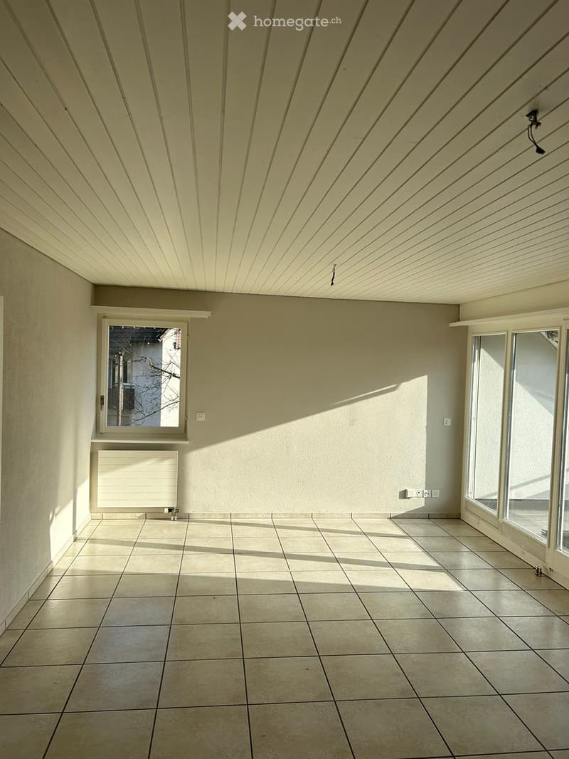 Empty room with tiled floor, light-colored walls, and large windows allowing natural light. The ceiling has a geometric pattern with recessed lighting.