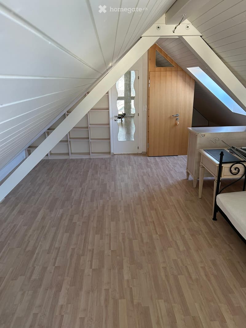 Attic room with wooden flooring, slanted ceilings, and skylight windows. Features a small table and chair near the window.