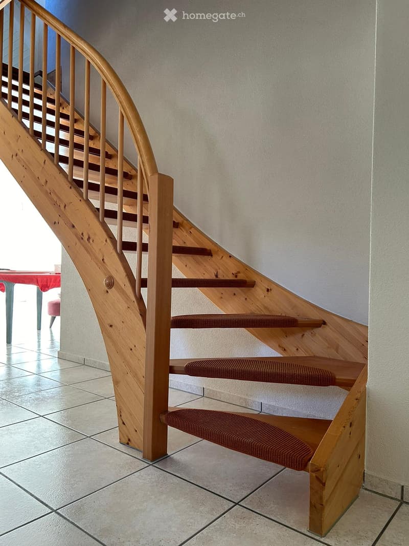 Wooden staircase with red carpeted steps and a wooden handrail, leading up to a higher level. The surrounding area includes tiled flooring and a glimpse of a red table and pink chair in the background.