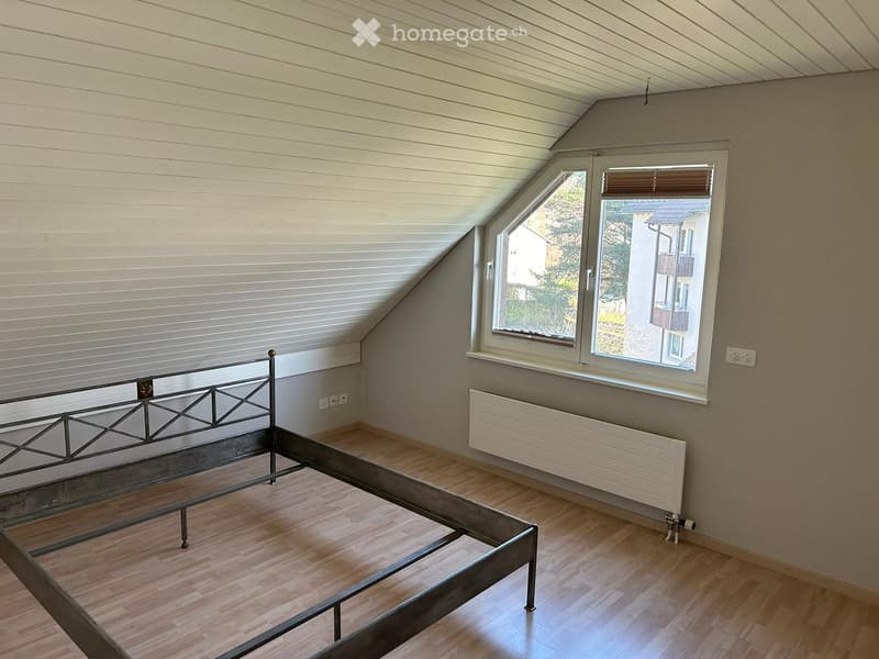 Empty attic bedroom with wooden flooring, slanted ceiling, and large windows offering a view of the outside. A metal bed frame is present but no other furniture or items.