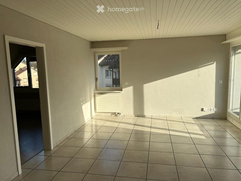 Empty room with tiled floor, white walls, and large windows allowing natural light. There is a door leading to another space and a radiator below one of the windows.