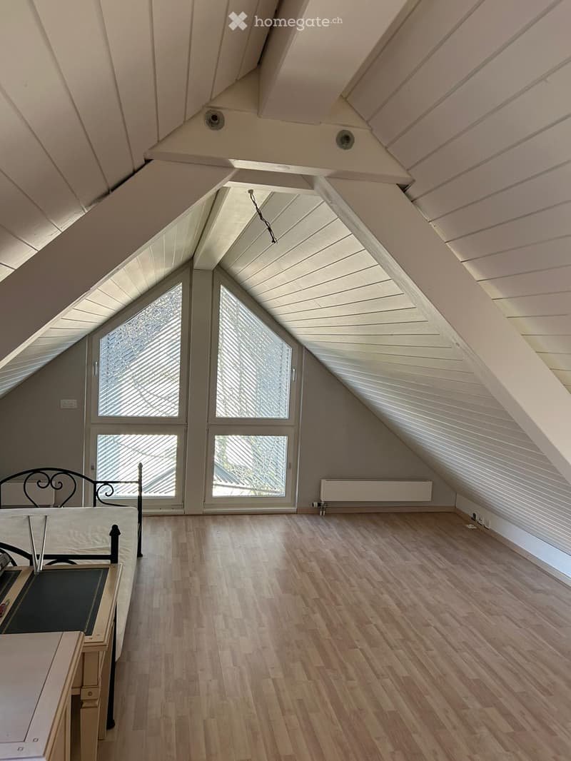 Empty attic room with wooden flooring, slanted white ceiling, large windows, and a radiator. No furniture or equipment present.