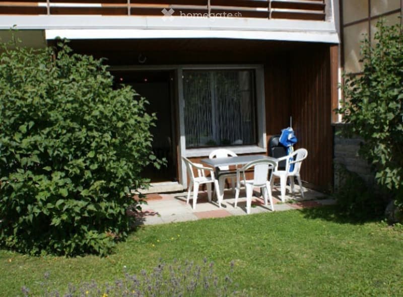 A small patio area with a table and chairs, located beneath a wooden balcony. The area is surrounded by green grass and bushes.