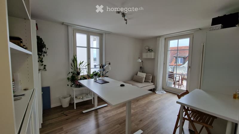 Bright, modern living space with wooden flooring, large windows, a sofa, a desk, and a balcony access.